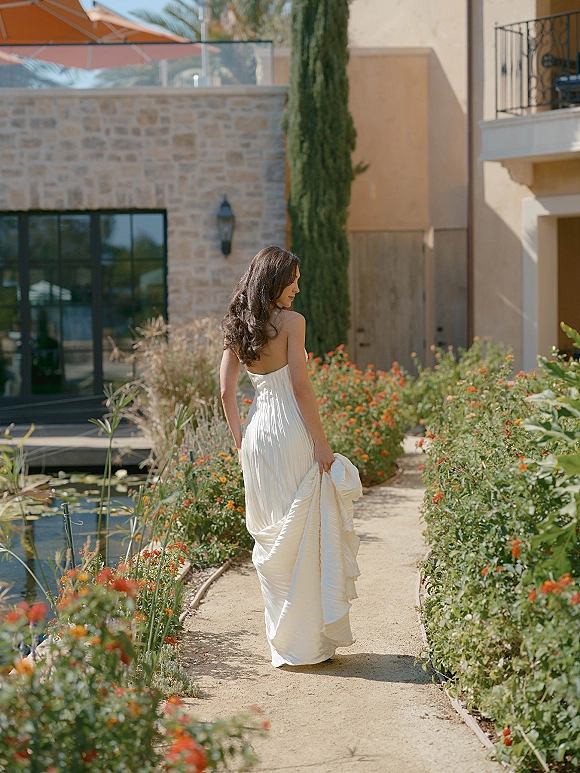 Bridal portrait of a bride walking away, holding her dress train in a strapless gown along a garden path by a pond and stone wall