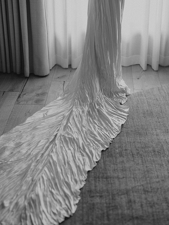 Wedding dress train in satin wedding dress fabric spread on a wood floor by window light, framed by curtains and an area rug