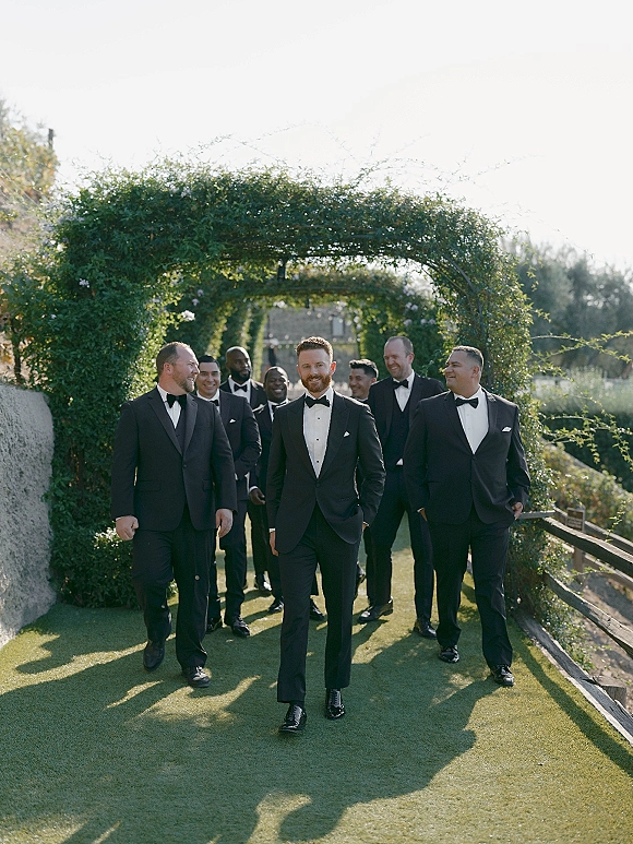 Groomsmen portrait of the groom with groomsmen in black tuxedos and bow ties, walking under a garden archway in sunlight