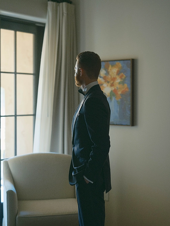 Groom portrait in tuxedo with a bow tie and pocket square, hands in pockets by a window with curtains in a hotel room