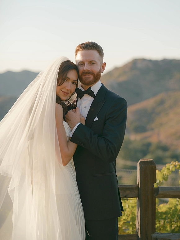 Couple portrait of bride and groom leaning together, her bridal veil flowing beside a wooden fence with mountains at sunset