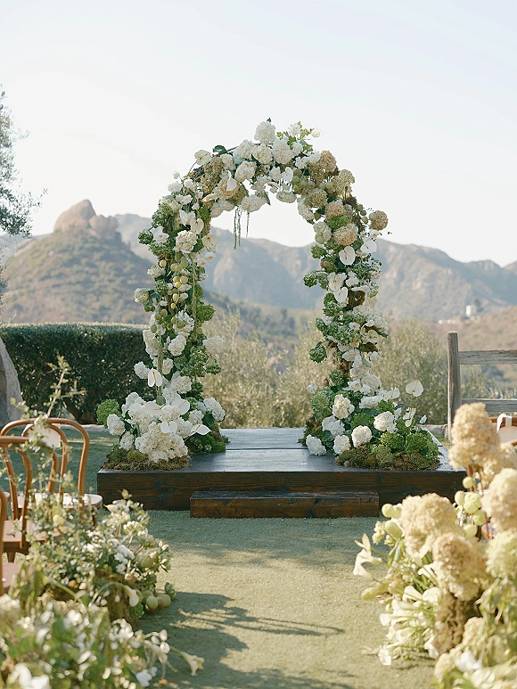 Wedding ceremony arch with a floral ceremony arch of white blooms and greenery on a wooden platform, framed by mountain views and lawn aisle florals