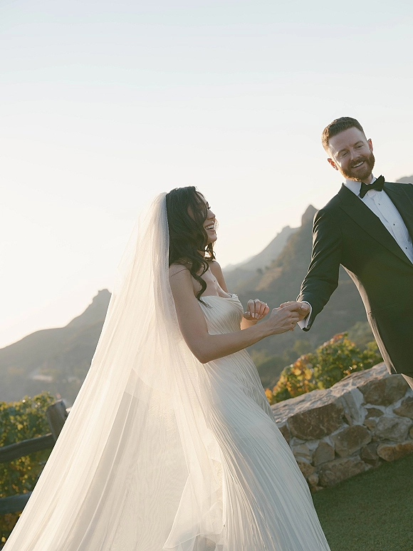 Couple portrait of bride and groom holding hands, her long veil blowing as they laugh by a stone wall with mountains and sky behind
