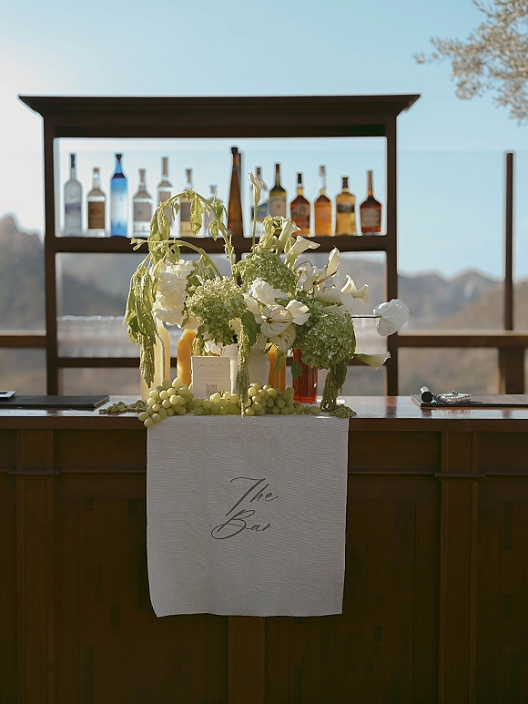 Wedding bar setup with liquor bottles on a shelf, green and white flowers, and a wooden counter on an outdoor deck with mountain view