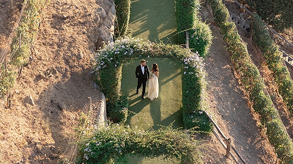 Couple portrait of bride and groom holding hands under a hedge arch with flowering vines, vineyard rows stretching behind them