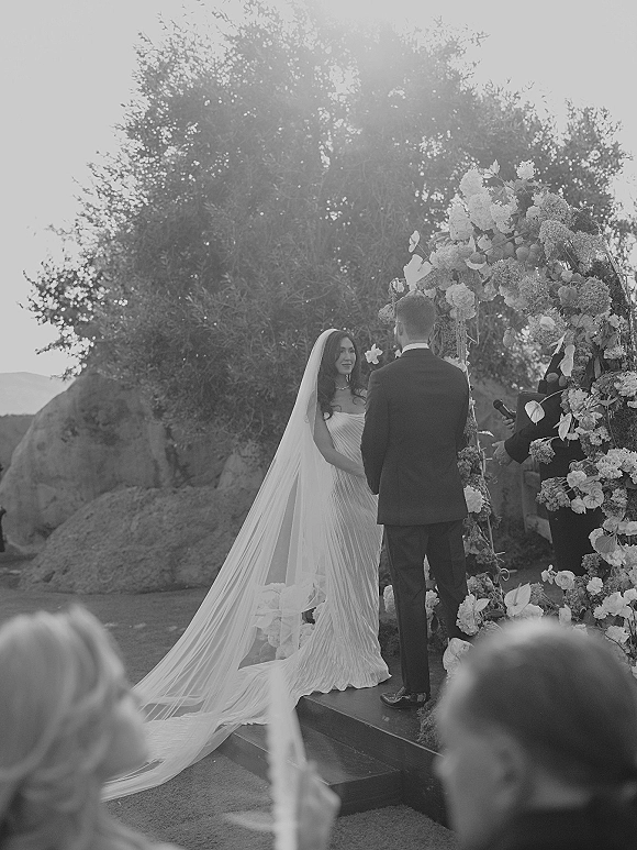 Wedding vows as bride and groom hold hands under a floral arch, veil flowing behind her while officiant speaks at outdoor altar steps