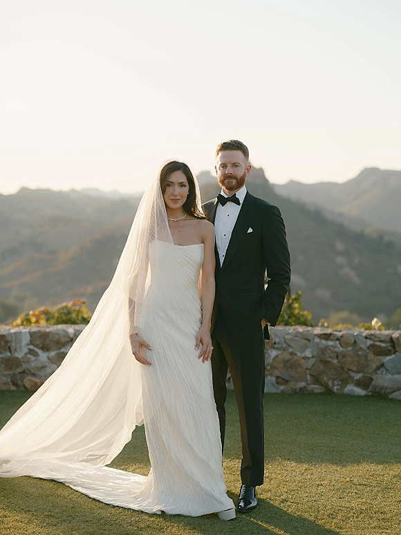 Couple portrait of bride in strapless wedding dress with cathedral veil and groom in tuxedo, posing by stone wall at sunset mountains