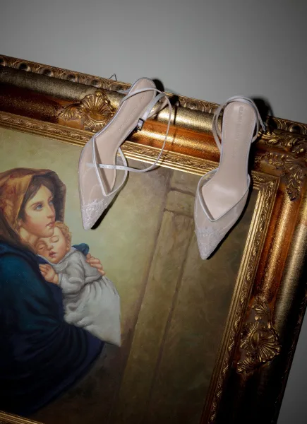 Bridal shoes with wedding heels detail, white strappy pointed-toe heels with lace and ankle straps resting on an ornate gold frame against a gray wall