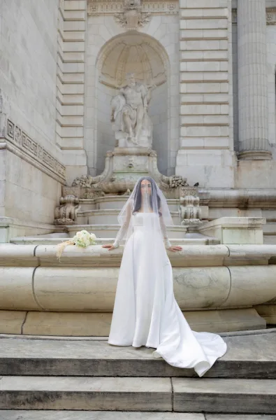 Bridal portrait of a bride in a strapless wedding dress with veil, holding a white rose bouquet by a stone fountain and columns