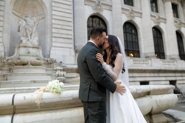 Wedding kiss portrait of bride and groom kissing by a stone balustrade, bride in long veil holding a simple white bouquet before statues and fountain