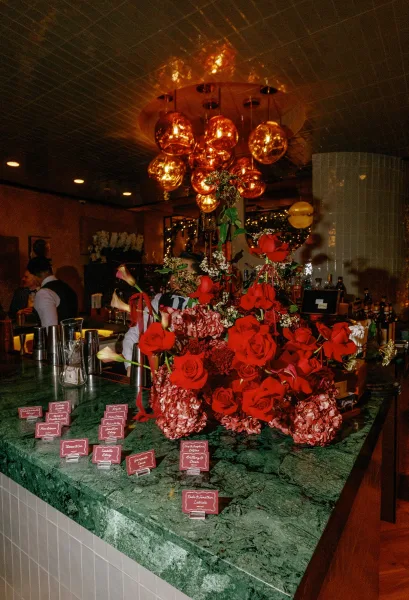 Escort card display with wedding escort cards on acrylic holders, framed by red roses and pink hydrangeas on a green marble bar top under copper pendant lights