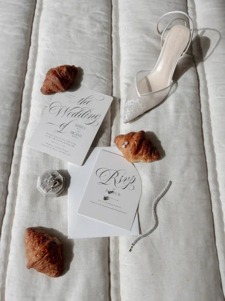 Wedding invitation suite with calligraphy wedding invitation, RSVP card, rings and lace heel on white quilted bedspread in soft light