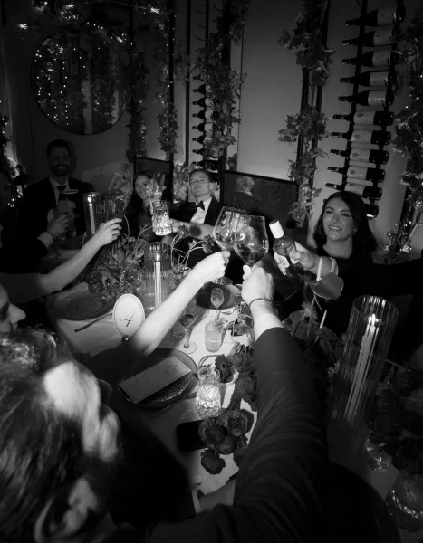 Wedding toast as guests clink wine glasses over a candlelit reception table with floral centerpiece and string lights in a restaurant interior