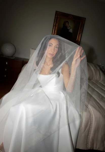 Bridal portrait of a bride under veil, seated on a bed in a bedroom, wearing a satin wedding dress and necklace under soft flash
