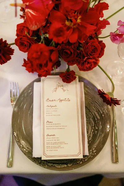 Reception tablescape with wedding place setting, black charger plate and menu card on napkin beside red floral centerpiece on white tablecloth