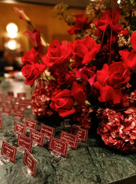 Escort card table with wedding escort cards in acrylic holders and red roses on a green tablecloth under warm bistro lights in a dim venue