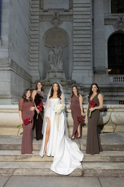Bride with bridesmaids posing on stone steps outside a classical building, bride in veil and slit gown holding a white bouquet, maids with red roses