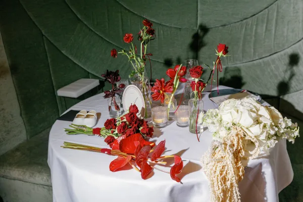 Wedding reception tablescape with round table centerpiece of red flowers, hydrangeas, candles, menus, bridal shoes, and pearl veil on green banquette