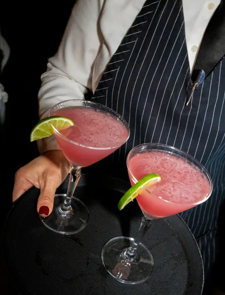 Signature cocktails on a serving tray with cocktail glasses and lime wedges, held by a bartender in vest and lapel pin in a dark indoor setting