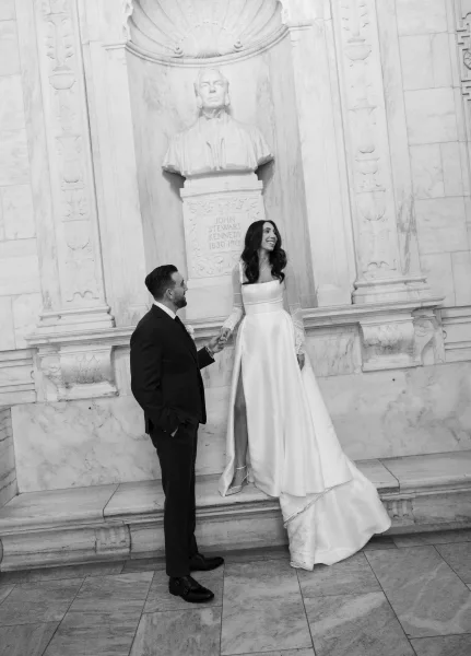 Couple portrait of bride and groom holding hands, groom watching her as she looks away by a marble wall with statue and columns.