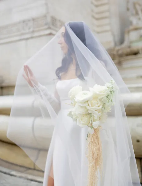 Bridal portrait of a bride with veil in a strapless gown, holding a white rose and greenery bouquet on stone steps by a building railing