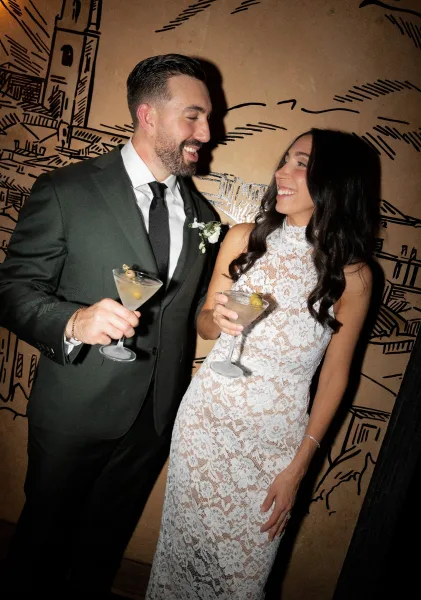 Couple portrait of bride and groom with drinks, toasting martinis with olive garnish in front of an illustrated mural wall backdrop