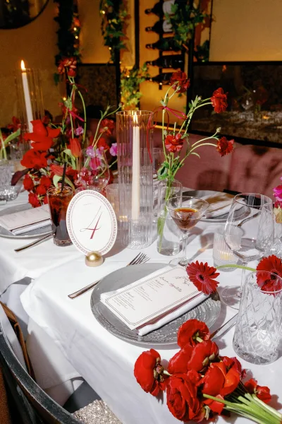 Reception tablescape wedding table setting with red flowers in bud vases, taper candles, and mix-and-match glassware by banquette seating and mirror