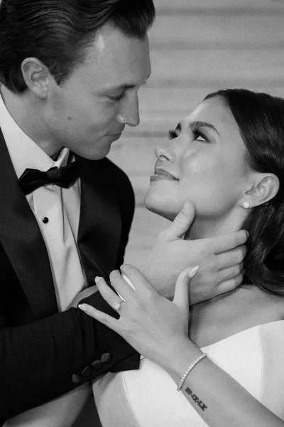 Wedding couple portrait in black and white, groom in tuxedo holding bride’s face as they lean forehead to forehead against a neutral wall