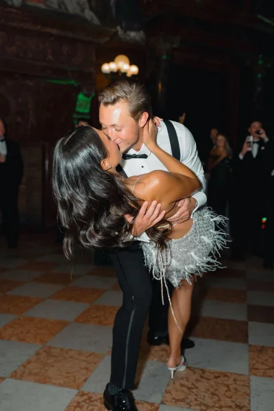 First dance moment as groom in suspenders dips bride in a feather mini dress for a kiss on a chandelier-lit dance floor amid guests in a dark reception room