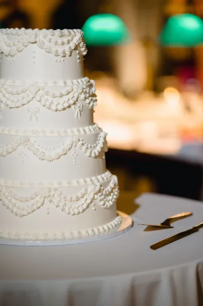 Wedding cake with piped frosting on a stand, set with cake knife and server on a round table under bokeh lights and green pendant lamps