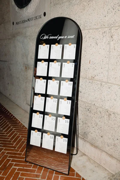 Wedding seating chart on an arched mirror with calligraphy cards and gold wax seals on a black stand against a stone wall backdrop