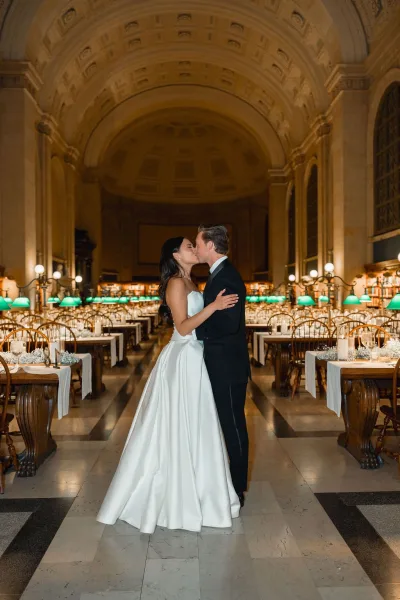 Wedding kiss portrait of bride and groom kissing, her strapless satin wedding dress and his tuxedo beneath globe lights in a grand library hall