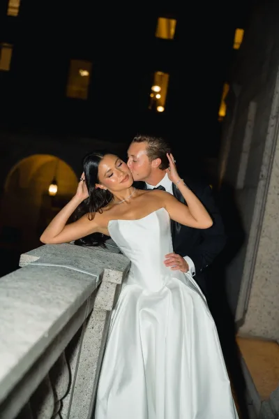 Wedding couple portrait of groom kissing bride in a strapless gown on a stone balcony, lantern lights glowing against lit windows at night