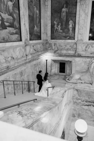 Couple portrait of bride and groom walking up a marble staircase, her wedding dress train flowing past murals and a statue in black and white