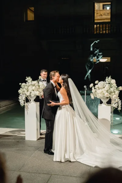 Wedding kiss as bride in a long veil and groom in tuxedo embrace by a reflecting pool fountain, framed by floral pedestals at night
