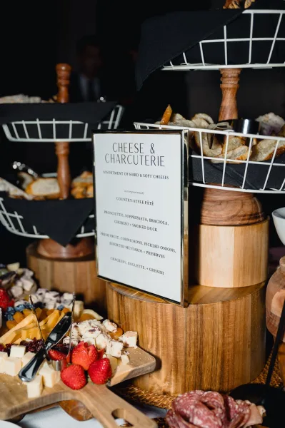 Charcuterie display with cheeses, cured meats, fruit, and breadsticks on wooden risers with a menu sign on a buffet table