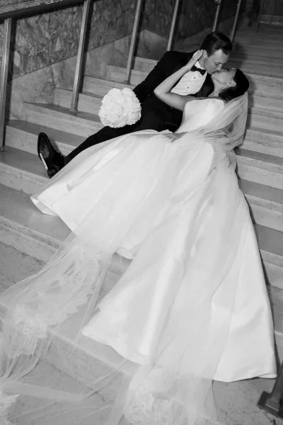 Wedding kiss portrait of bride and groom kiss in a dramatic dip on stone stairs, her long veil and white bouquet cascading beside a glass railing