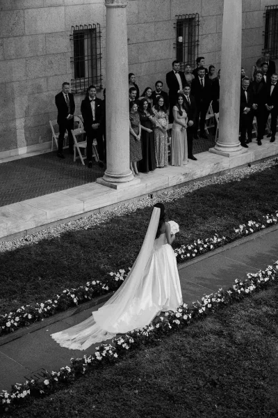 Wedding processional with bride walking down aisle from behind, cathedral veil and long train, bouquet in hand, guests by stone columns