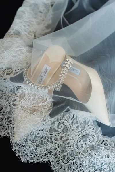 Wedding shoes with rhinestone strap heels resting on lace fabric and bridal veil in a flatlay on a dark surface, elegant getting-ready detail