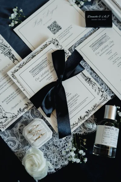 Invitation suite with black and white wedding invitations tied in a black satin ribbon, styled with rings and a white rose on a dark surface