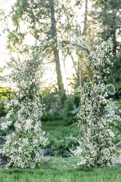 Wedding ceremony arch with white flowers and greenery accents on a wooden frame, set on a sunlit garden lawn with trees behind