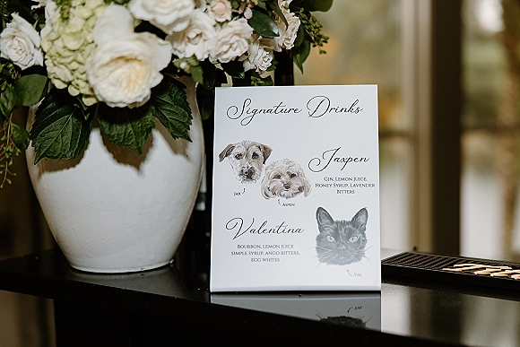 Signature drink sign and wedding signature drinks menu with calligraphy and watercolor pets beside white roses in a ceramic vase on black tabletop
