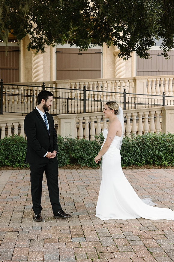First look moment as bride in strapless dress and veil faces groom in black suit by stone balustrade in brick courtyard.