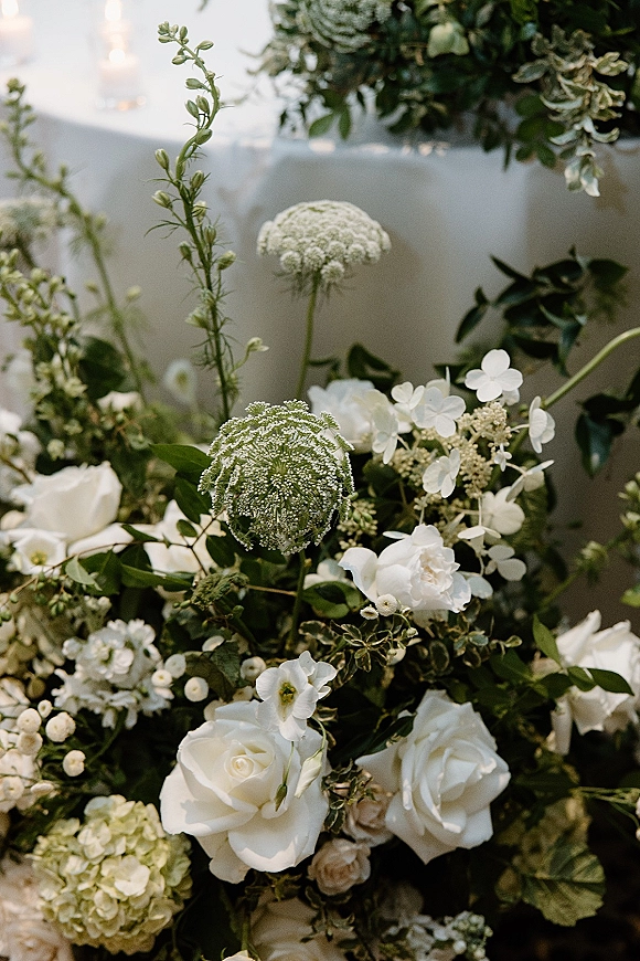 Wedding floral arrangement of white wedding flowers with roses, hydrangea, and greenery on a linen table with candles and bokeh lights