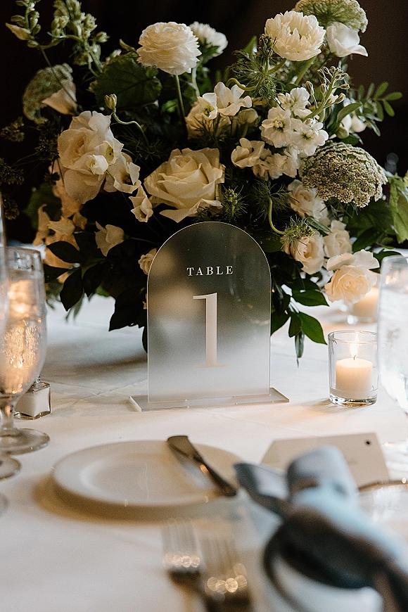 Reception tablescape with a wedding table centerpiece of white florals, arched table number sign, votive candles, and place setting in a dark room