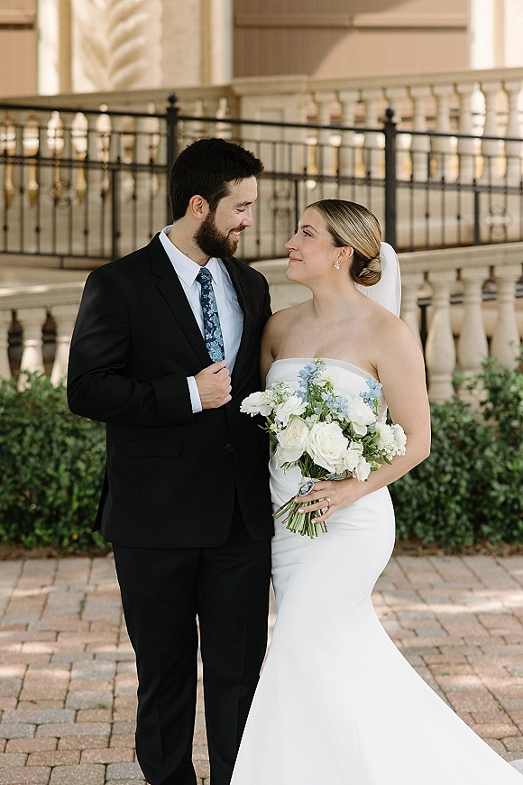 Couple portrait of bride and groom portrait looking at each other, bride holding bouquet of white roses with blue blooms by a stone facade balcony