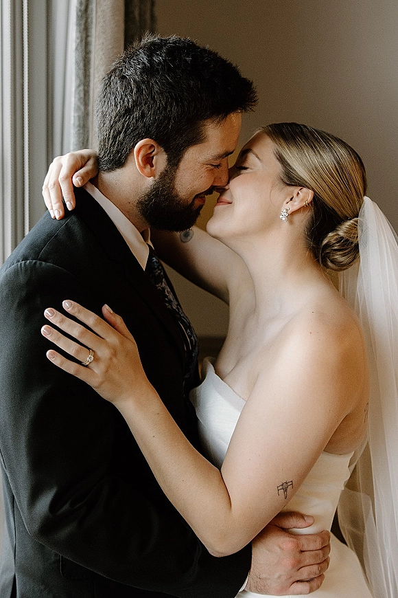 Wedding couple portrait of bride and groom embracing nose to nose, bridal veil framing her strapless dress in soft window light indoors