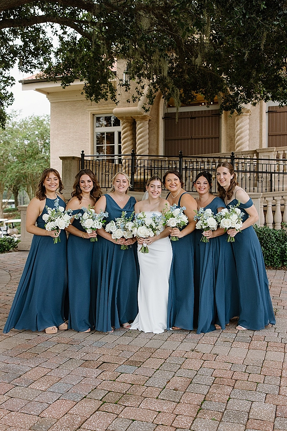 Bridesmaids portrait with bride with bridesmaids holding white rose and greenery bouquets in front of a villa facade and balcony railing