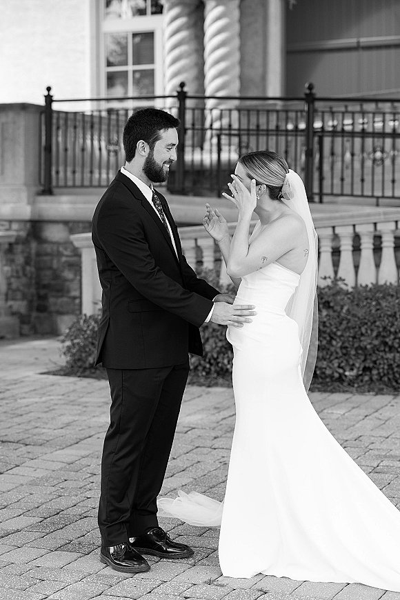 First look moment as bride covers her face in veil, holding hands with groom in suit on a stone patio by iron railing