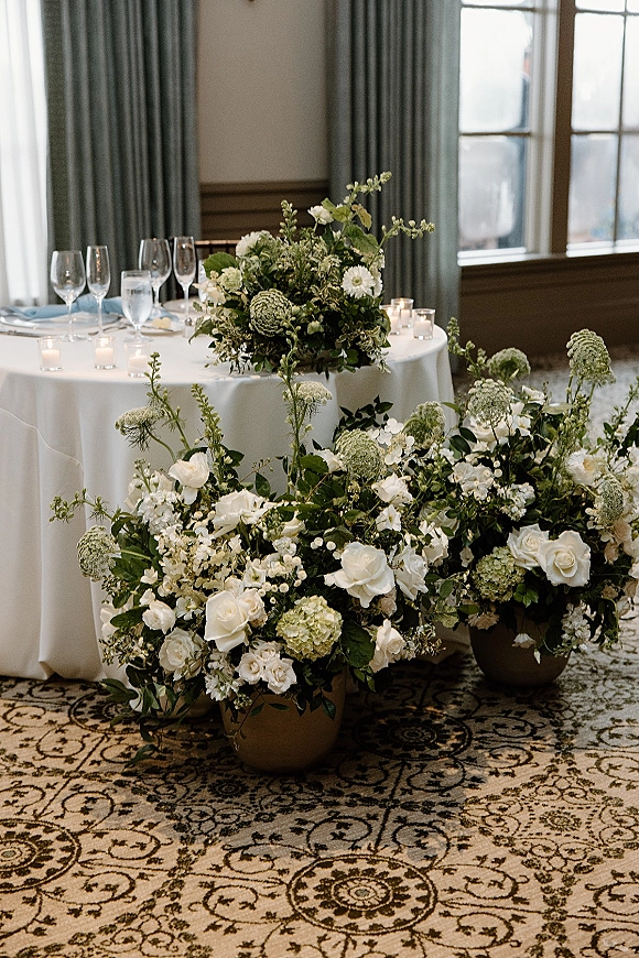 Reception tablescape with white and green centerpiece, bud vases and votive candles on a round table by large windows with curtains